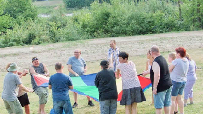 Gäste des Lebenshilfe-Sommerfestes spielen mit dem Fallschirm verschiedene Spiele. Zu sehen sind die Gäste, die einen bunten Fallschirm schwingen. 
