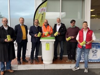 Sieben Männer präsentieren grüne Sammelbehälter vor einer Recyclingtonne.