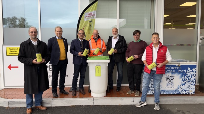 Sieben Männer präsentieren grüne Sammelbehälter vor einer Recyclingtonne.
