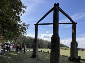 Große hölzerne Tor-Skulptur auf einer Wiese mit Besuchergruppe.