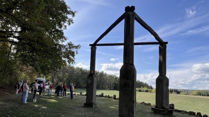 Große hölzerne Tor-Skulptur auf einer Wiese mit Besuchergruppe.