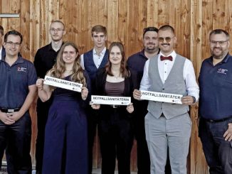 Neun Absolventen feiern ihre Zertifizierung als Notfallsanitäter vor Holzhintergrund.