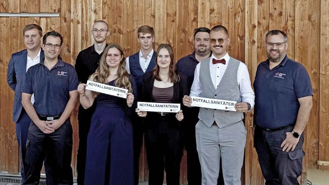 Neun Absolventen feiern ihre Zertifizierung als Notfallsanitäter vor Holzhintergrund.